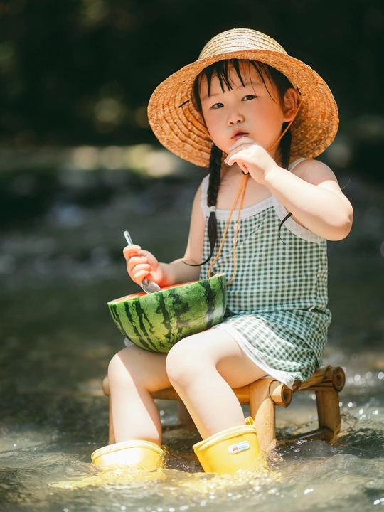一个戴草帽的小女孩坐在木制小溪上 手里拿着一个小西瓜在水里玩耍。