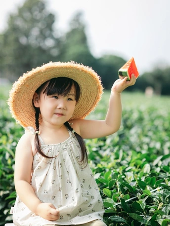 一个戴草帽的小女孩手里拿着一个草莓 她站在草莓植株的田地里 这是一个花园。