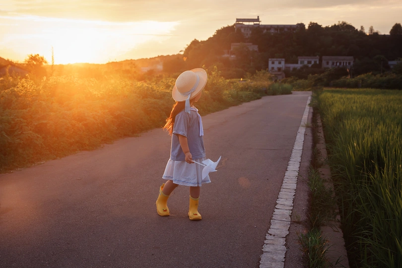 一个穿着白色连衣裙、黄色靴子和白色帽子的年轻女孩在日落时沿着道路走下去。