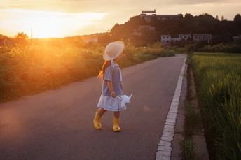 一个穿着白色连衣裙、黄色靴子和白色帽子的年轻女孩在日落时沿着道路走下去。