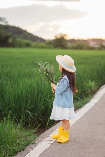 一个小女孩穿着黄靴子 手里拿着一个小植物和一把雨伞 站在一条路上。