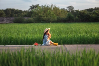 一个戴黄帽的小女孩坐在绿草地上 路边有一把黄色的伞。