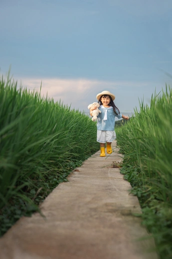 一个小女孩穿着裙子和小帽子站在一条小径上 这条小径位于高高的绿色草丛和草地上。