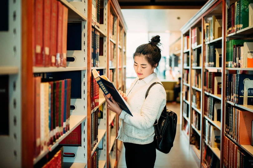 一个年轻女子站在图书馆的书架之间 正在阅读一本书。