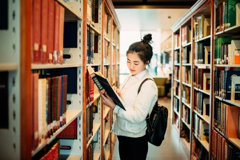 一个年轻女子站在图书馆的书架之间 正在阅读一本书。