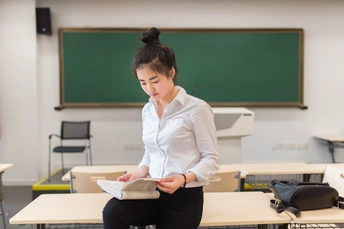 一个年轻女子坐在教室的桌子旁阅读一本书 一名女教师站在她身后。