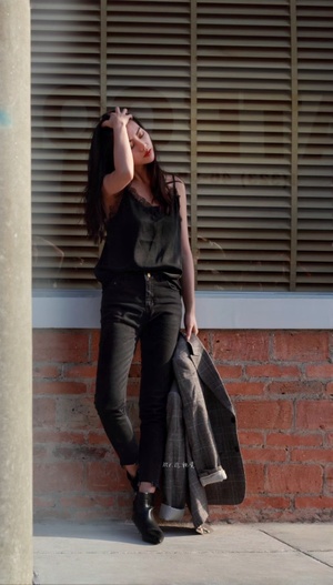 一位穿着全黑衣服的年轻女子靠在一栋建筑物的 sidewalk 上 紧靠着砖墙。