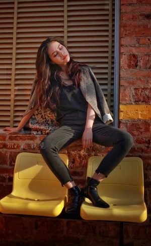 一个穿着黑色靴子的年轻女子坐在黄色长凳上 面对砖墙。