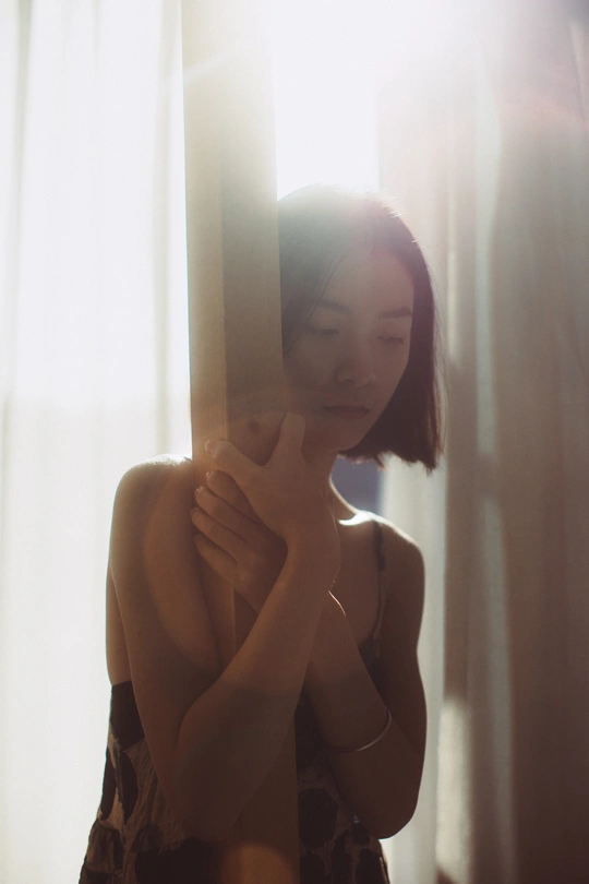 一位年轻女子举起双手捂住脸庞 正对着有薄纱窗帘遮盖的镜子凝视。