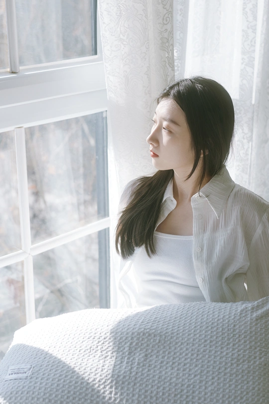 一个年轻女子穿着白色衬衫坐在床上 凝视着窗户。