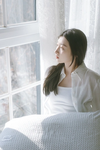 一个年轻女子穿着白色衬衫坐在床上 凝视着窗户。
