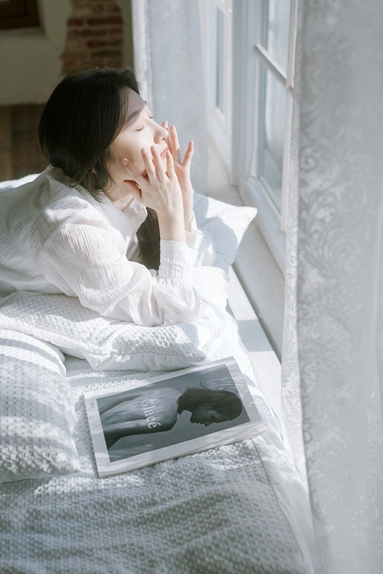 一个年轻女子坐在床上 双手捂住脸 旁边放着一本书 正在凝视窗外。