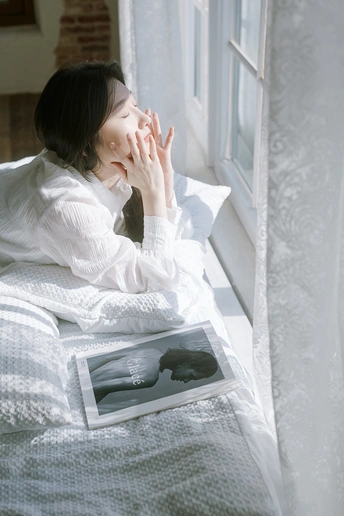 一个年轻女子坐在床上 双手捂住脸 旁边放着一本书 正在凝视窗外。