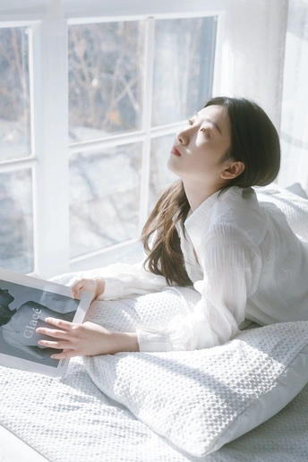 一个年轻女子坐在白色床上阅读一本书 凝视着窗外。