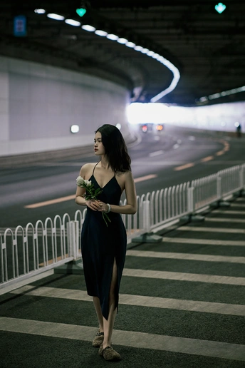 一个穿着黑色连衣裙的年轻女子手拿玫瑰和伞在夜晚走在街上