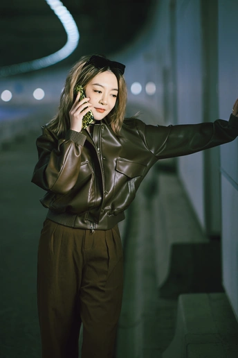 一个披着夹克的女青年在夜晚倚靠在墙上 用手机对着耳朵讲话。