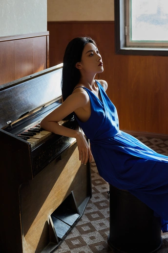 一位穿着蓝色连衣裙的年轻女子 倚靠在一架钢琴前 面对窗户。