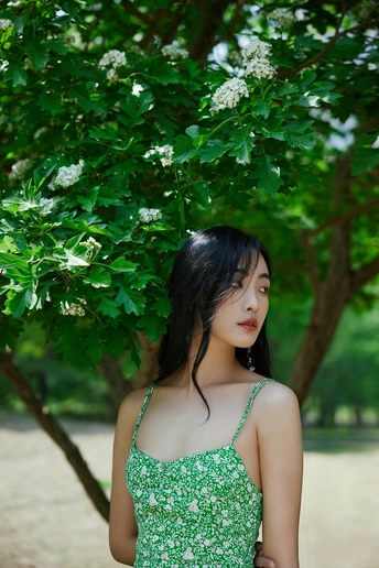 一位穿着绿色连衣裙的年轻女子站在公园里 周围有树木和花。
