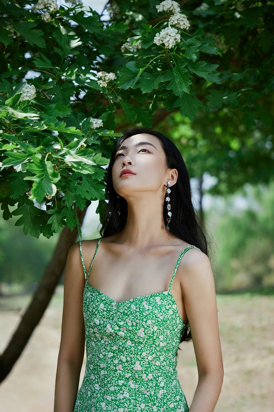 一位穿着绿色连衣裙的年轻女子站在一棵树下 花丛中。