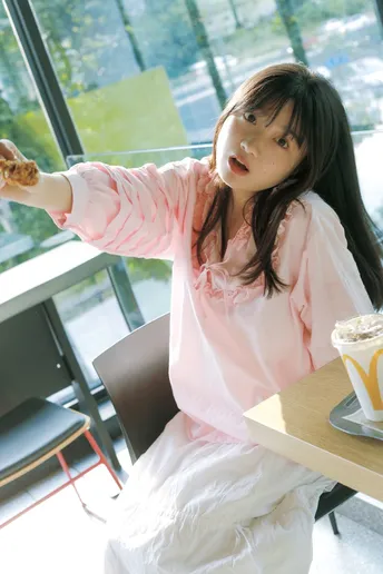 一个穿粉红色衣服的年轻女孩坐在桌子旁 喝着一杯咖啡和一个甜甜圈 正在吃早餐。