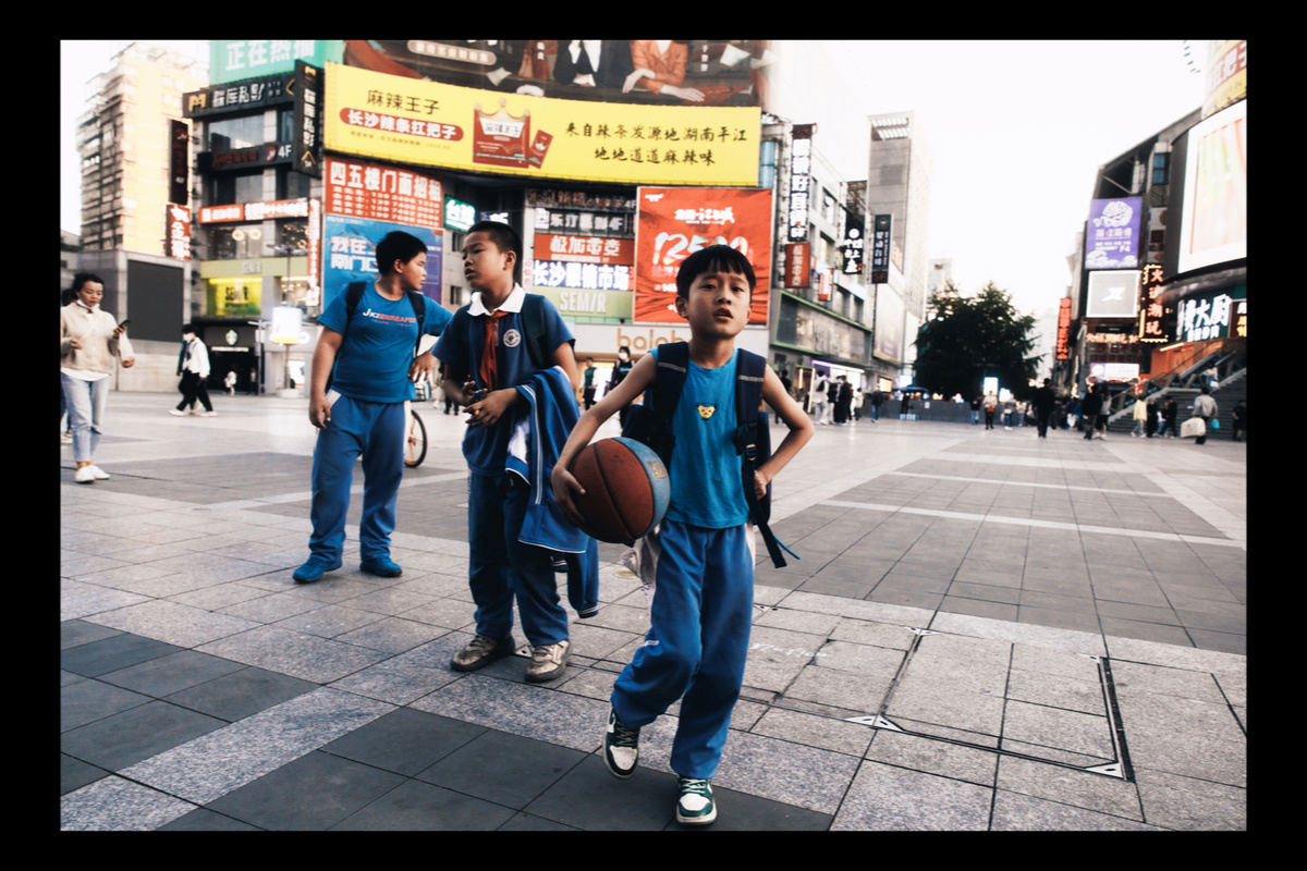 一个小男孩拿着一个篮球站在人行道上 看着经过城市的人们。