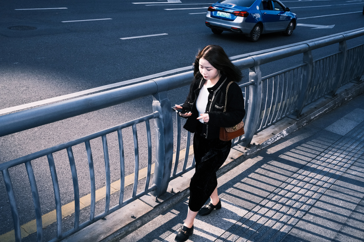 一位年轻女子站在人行道上 桥上正在看手机 桥下是繁忙的街道。