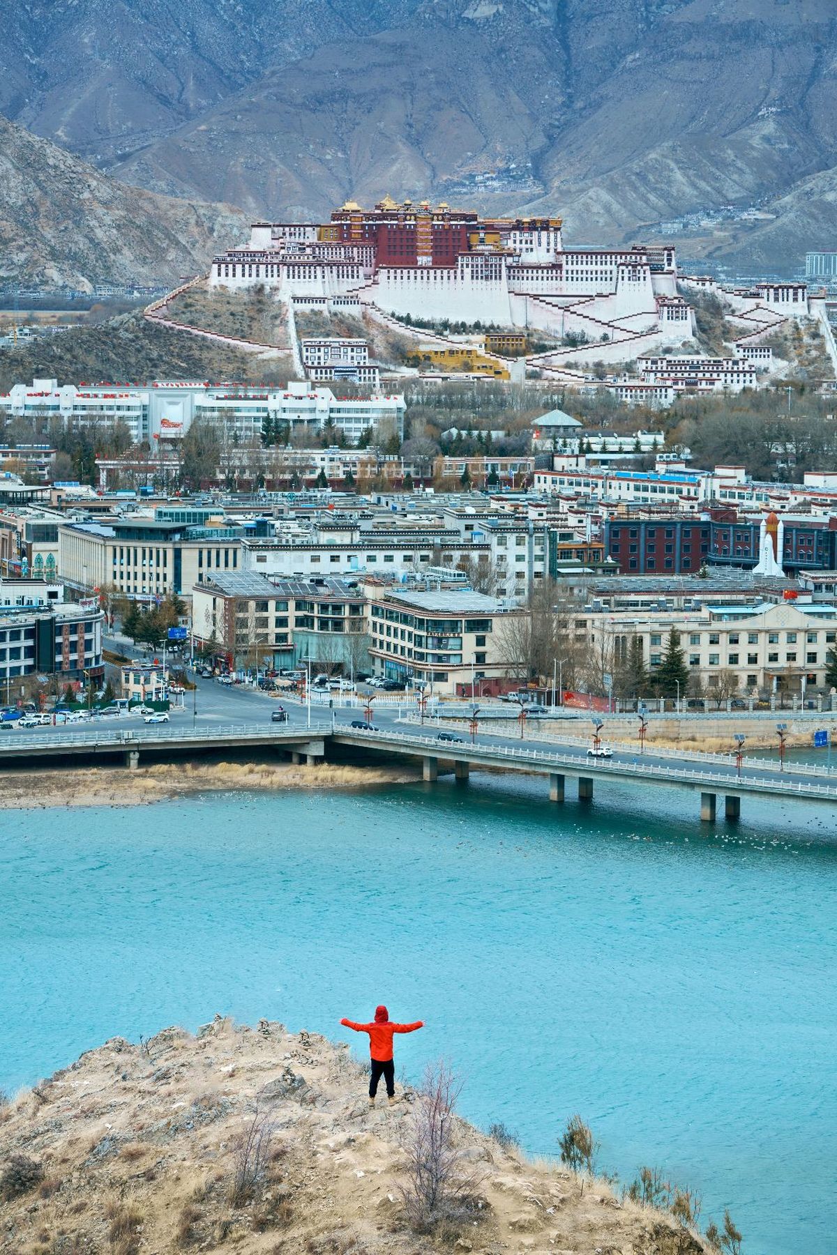 一个穿红夹克的男人站在俯瞰城市和蓝色海水的山丘上