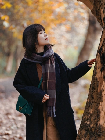 一位年轻女子穿着一件外套和围巾 靠在一棵树旁