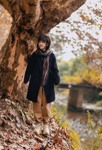 一位年轻女子站在一棵有落叶的树旁边 穿着一件外套 还有一个小男孩。