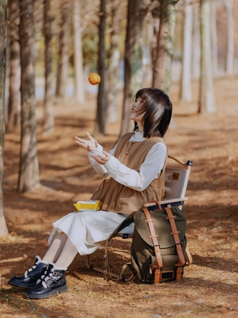 一位年轻女子坐在森林中的一把椅子上 背着背包 拿着笔记本电脑 手里拿了一个橙子。
