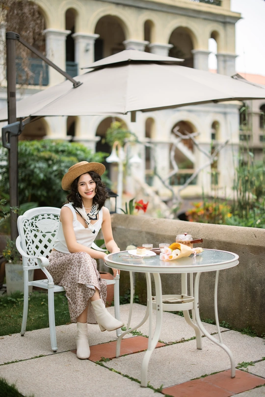 一个戴着帽子的年轻女人坐在院子里的白色椅子上 手里拿着雨伞和一张白色的小桌子