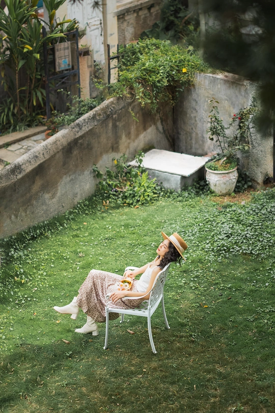 一个穿着连衣裙、戴着帽子的女人坐在院子里的椅子上 一只猫躺在草地上 一条狗躺在背景中