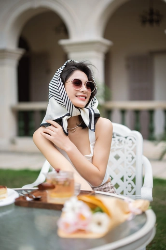 一个戴着墨镜和围巾的年轻女子拿着一盘食物坐在外面的桌子旁