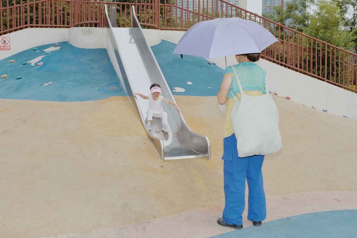 一位女士拿着一把雨伞站在水上乐园的滑梯旁边 旁边有一个孩子。