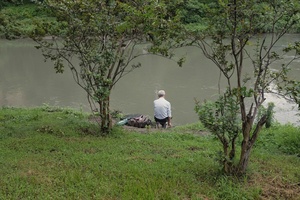一个人坐在流入水体的河流的河岸上 对岸有一棵树和一个人。