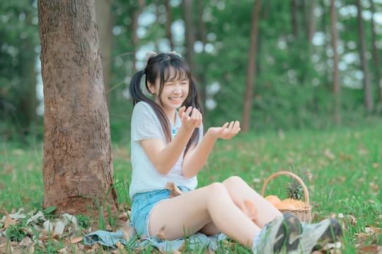 一位年轻女子坐在森林里一棵树旁的草地上 面前放着一篮树叶