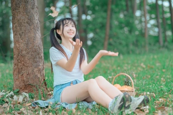 年轻漂亮的女人坐在树林里一棵树旁的草地上 面前拿着一个篮子 挥手