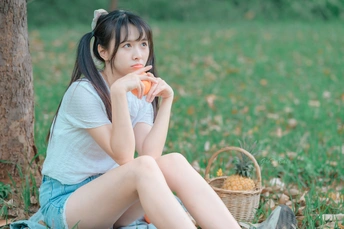 一个年轻的女人拿着一篮胡萝卜坐在树下的草地上吃橘子