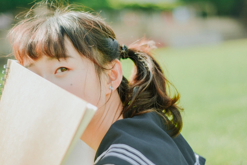 一个小女孩坐在公园的草地上 手里拿着一本书 挡在脸前。