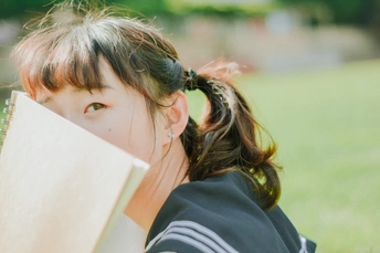 一个小女孩坐在公园的草地上 手里拿着一本书 挡在脸前。