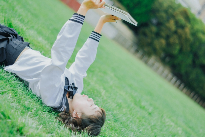 一个年轻女子躺在绿草地上 头上放着一本书和一台笔记本电脑。