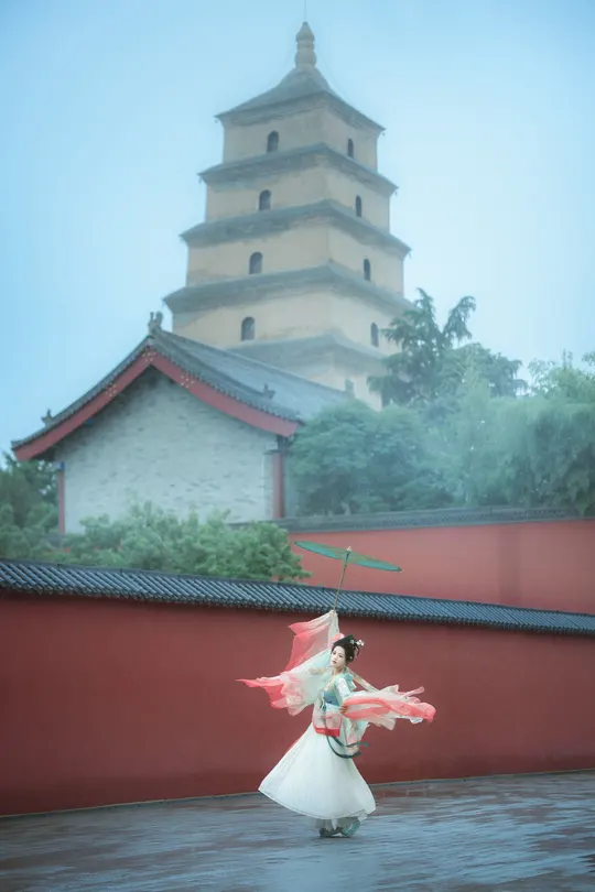 一个穿白裙子的女孩在雨中站在红色建筑前 手里拿着一把雨伞。