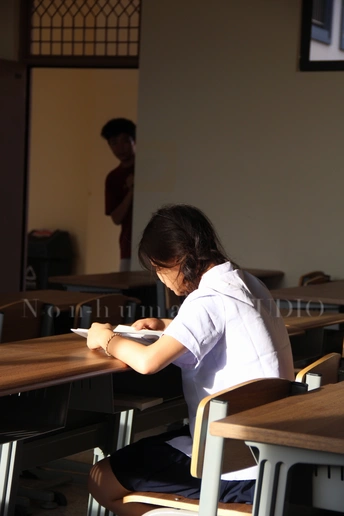 一个年轻女孩坐在教室里的一张桌子旁 周围是空椅子 正在写一本书。