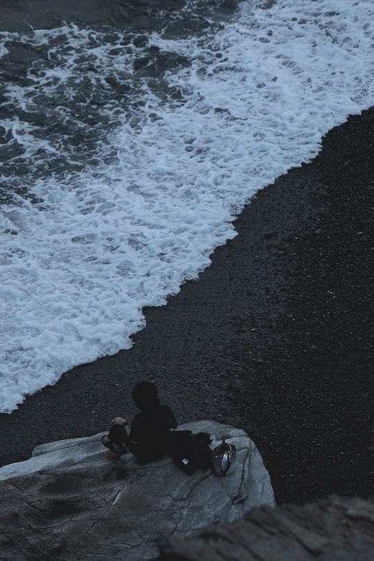 一个男孩坐在黑沙滩的边缘一块岩石上 海浪拍打着海洋的岸边。