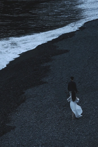 一个穿白裙子的男人沿着一个黑沙滩的海岸走