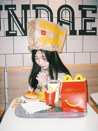 一个戴纸袋的年轻女子坐在桌子前 面前放着汉堡和饮料。