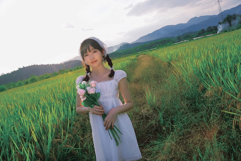一个小女孩穿着白色裙子 手里拿着花站在绿色的田野里。