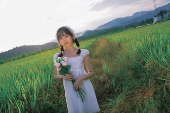 一个小女孩穿着白色裙子 手里拿着花站在绿色的田野里。