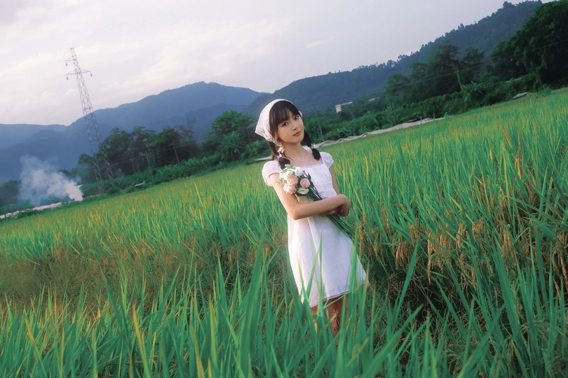 一个小女孩穿着白色裙子 手里拿着一朵花站在一片高草地上。