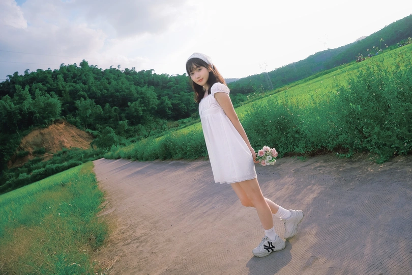 一个穿着白色连衣裙的年轻女子站在一条路的中间 靠近一片花田。